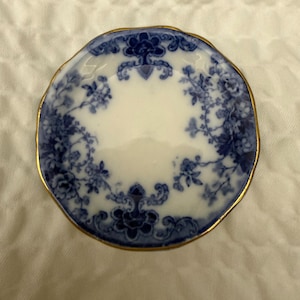 May include: A small, round ceramic dish featuring a white center and a blue floral pattern around the rim. The edge of the dish is finished with a thin gold-colored trim, and the overall design includes intricate blue botanical motifs.