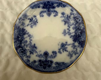 May include: A small, round ceramic dish featuring a white center and a blue floral pattern around the rim. The edge of the dish is finished with a thin gold-colored trim, and the overall design includes intricate blue botanical motifs.