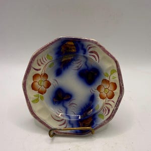 May include: An octagonal ceramic bowl featuring a cream-colored base with hand-painted orange flowers, green leaves, and deep blue abstract cloud-like patterns. The rim is accented with a thin, textured purple border, and the surface has a visible crackle glaze finish.