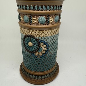 May include: A decorative ceramic vase with a cylindrical shape. The vase features a mosaic-like pattern in shades of teal, beige, and brown. The design includes floral and geometric motifs, with a textured surface. The vase has a wide base and a slightly flared top.