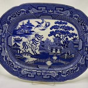 May include: An oval ceramic platter featuring a blue and white willow pattern. The design depicts a landscape with pagodas, a bridge, a boat, and two birds in flight, surrounded by a wide border of geometric and floral motifs on a white base.
