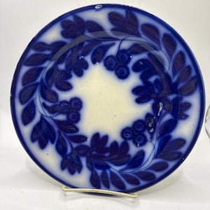 May include: A round ceramic plate featuring a deep blue transferware pattern of leafy branches and clusters of berries arranged in a circular wreath design against a creamy white background, with a solid blue rim and a smooth glazed finish.