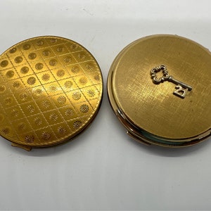 May include: Two gold-colored compact mirrors. One mirror has a floral pattern and the other has a key with the number 21 on it.