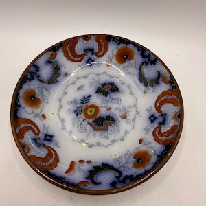May include: A small, round, antique plate with a white center and a brown rim. The plate features a floral design with orange and blue accents. The design includes flowers and ornate patterns, creating a vintage aesthetic.