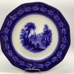 May include: A blue and white porcelain plate with a decorative border and a central image of a landscape with a building and trees.