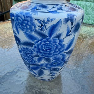 May include: A white ceramic vase with a blue floral design. The vase features large blue flowers and leaves, with a gold rim at the top. The vase is a decorative item.