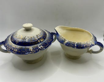 May include: A vintage cream and sugar set. The sugar bowl has a lid and handles, and the creamer has a spout and handle. Both pieces are white with blue and gold floral detailing.