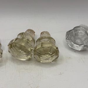 May include: A collection of eight clear glass cabinet knobs featuring a faceted, multi-sided design. The knobs are arranged in three small groups, showcasing a transparent, reflective surface with a standard threaded metal base for installation.