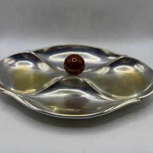 May include: A silver-toned, three-section serving dish with a central, round, brown accent. The dish has a leaf-like shape with curved edges and a reflective surface. A decorative piece for serving snacks or appetizers.