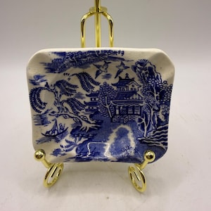 May include: A square ceramic trinket dish featuring a blue and white willow pattern illustration of a pagoda, willow trees, and birds on a river. The dish is displayed on a gold-toned metal wire stand with a curved base and a vertical support.