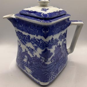 May include: A square-shaped ceramic teapot featuring a classic blue and white willow pattern. The design depicts a landscape with pagodas, bridges, and trees. It includes a matching lid with a small knob handle, a curved spout, and a side handle.