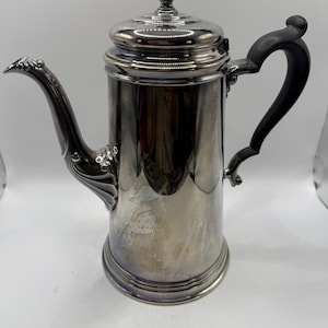 May include: A silver-colored coffee pot with a black handle and a decorative spout. The pot has a lid with a detailed finial. The design is vintage and elegant, suitable for serving coffee or tea.