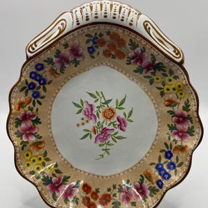 May include: A scalloped ceramic serving dish featuring a central floral bouquet and a wide tan border decorated with colorful flowers in shades of pink, orange, yellow, and blue. The rim is accented with white and gold detailing along the edges.