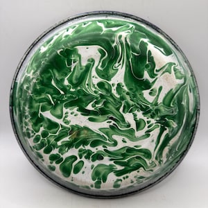 May include: Round enamelware plate with a green and white marbled pattern. The plate has a dark gray rim and a white interior with a swirling design. The plate is likely vintage and suitable for serving food.
