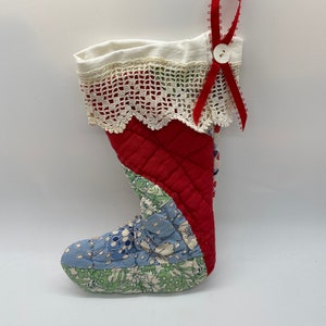 May include: A quilted Christmas stocking with a red, white, and blue patchwork design. The stocking features a red quilted body, a white lace trim, and a red ribbon loop with a white button. The toe is blue with white floral patterns.