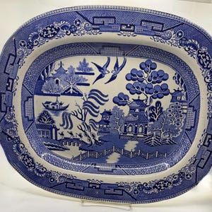 May include: An oval ceramic serving platter featuring a blue and white willow pattern. The design depicts a landscape with pagodas, a bridge, willow trees, and two birds in flight, surrounded by a decorative geometric border.