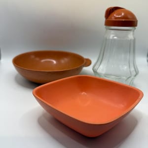 May include: An assortment of kitchenware, including a clear glass salt shaker with an orange lid, a brown bowl with a handle, and an orange square bowl. The items are arranged on a white surface.