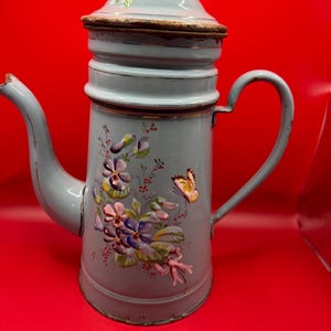 May include: A light blue enamel coffee pot with a lid and handle. The pot features hand-painted floral and butterfly designs in shades of purple, pink, and yellow. The pot is approximately 10 inches tall.