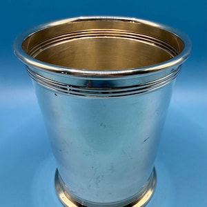 May include: A silver-colored metal tumbler with a wide, cylindrical shape. The tumbler has a polished finish with horizontal bands near the top and base. The interior has a gold-colored finish.