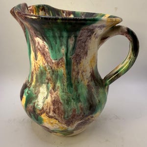May include: A ceramic pitcher with a unique, marbled design. The pitcher features a handle and a ruffled rim. The color palette includes green, brown, yellow, and cream, creating a swirled effect. The pitcher is a decorative item.
