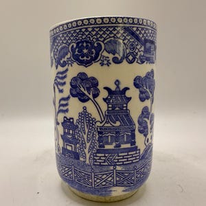May include: A cylindrical ceramic mug with a blue and white Willow pattern design. The pattern features a pagoda, trees, and a fence. The mug has a cream-colored base and a blue rim.