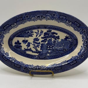 May include: An oval ceramic serving platter featuring a classic blue and white willow pattern. The design depicts a landscape with pagodas, willow trees, and birds in flight, surrounded by a decorative geometric border on a cream-colored background.