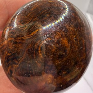 Pode incluir: Uma pedra circular polida com uma superf&iacute;cie lisa e brilhante. A pedra apresenta um padr&atilde;o rodopiante e marmoreado em tons profundos de castanho escuro, mogno e preto, com estrias cor de &acirc;mbar criando uma textura org&acirc;nica complexa.