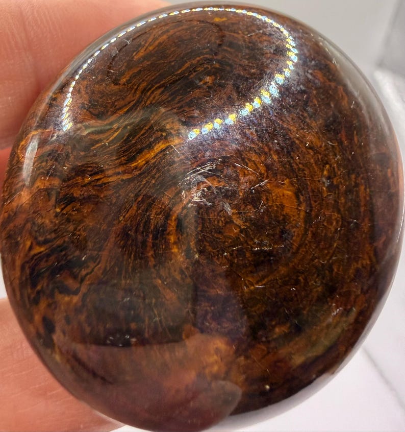 Pode incluir: Uma pedra circular polida com uma superf&iacute;cie lisa e brilhante. A pedra apresenta um padr&atilde;o rodopiante e marmoreado em tons profundos de castanho escuro, mogno e preto, com estrias cor de &acirc;mbar criando uma textura org&acirc;nica complexa.