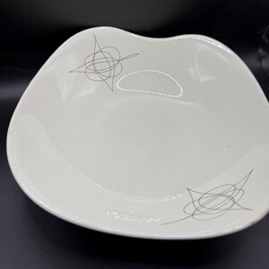 May include: A white, mid-century modern serving bowl with a unique, wavy shape. The bowl features a gray, abstract atomic design on the surface. The bowl is made of ceramic and is ideal for serving snacks or small dishes.