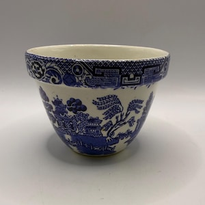 May include: A ceramic bowl featuring a blue and white willow pattern. The exterior displays detailed illustrations of traditional pagodas, willow trees, and a bridge. The wide, flared rim is decorated with a geometric border pattern in blue.