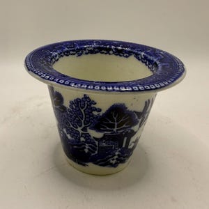 May include: A blue and white ceramic planter with a flared rim. The planter features a classic Willow pattern design, depicting a landscape scene with trees, a bridge, and a building. The rim has a detailed border.