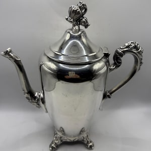 May include: A silver-colored teapot with ornate detailing. The teapot features a curved spout, a decorative handle, and a lid topped with a floral design. The teapot sits on three decorative feet.