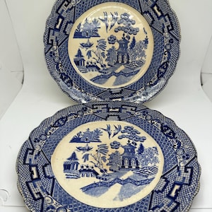 May include: Two blue and white ceramic plates with a willow pattern design. The plates have a scalloped edge and a blue and white geometric pattern around the rim.