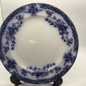 May include: A white ceramic plate with a blue and pink floral pattern around the rim. The plate has a gold trim.