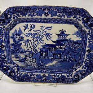 May include: An octagonal ceramic serving platter featuring a blue and white willow pattern. The design depicts a traditional landscape with pagodas, a willow tree, two birds in flight, a small boat on water, and a bridge, all framed by a decorative border.