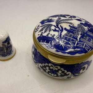 May include: A white porcelain thimble and a matching round trinket box, both decorated with a blue willow pattern. The trinket box features a gold-toned metal rim and a hinged lid, while the thimble has a gold-toned band at the base.