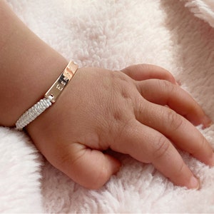 Op de afbeelding: Een wit gevlochten armband met een gouden naamplaatje waarop "Esme" staat.