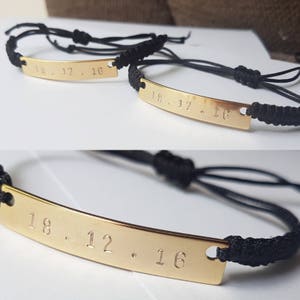 K&ouml;nnte beinhalten: Zwei schwarze Armb&auml;nder aus Schnur mit goldenen Metallplatten, die mit dem Datum "18.12.16" graviert sind.