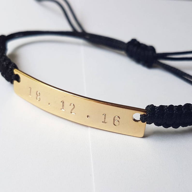K&ouml;nnte beinhalten: Ein schwarzes Kordelarmband mit einem goldenen Barren-Anh&auml;nger, der mit dem Datum "18.12.16" graviert ist.