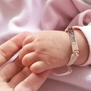 May include: A white braided bracelet with a rose gold nameplate that says "Brielle" with a heart symbol. The bracelet is on a baby's wrist.