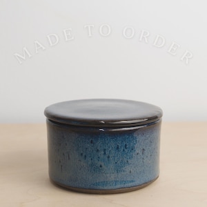 May include: A handmade, ceramic jar with a lid. The jar is cylindrical with a dark blue glaze and a speckled texture. The lid is a darker shade of blue. The words "MADE TO ORDER" are arched above the jar.