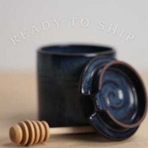 May include: A ceramic honey pot with a dark blue glaze, a matching lid, and a wooden honey dipper. The lid is partially open, revealing the interior. The pot and lid have a textured appearance. The wooden honey dipper is light brown.