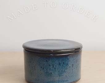 May include: A cylindrical ceramic container with a dark blue and black mottled glaze and a matching flat lid. The text MADE TO ORDER is visible in the upper portion of the image. The container has a smooth, glossy finish and a straight-sided body.