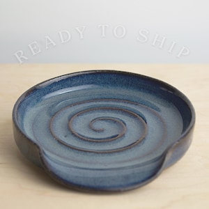 May include: A blue ceramic spoon rest with a spiral design in the center. The spoon rest has a dark rim and a curved edge for holding utensils. The text "READY TO SHIP" is visible above the spoon rest.