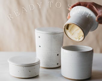 May include: A set of white ceramic butter crocks with dark speckles. One crock is covered with a lid, one is open, and one is held upside down showing butter inside. The text READY TO SHIP is visible in the background of this kitchenware set.