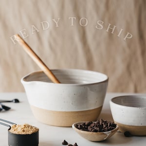 May include: A white and tan ceramic mixing bowl with a wooden spoon inside, alongside a small bowl of chocolate chips and a measuring cup of brown sugar. The words "READY TO SHIP" are arched above the bowl.