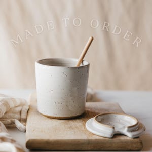 May include: A speckled white ceramic cup with a wooden stirring stick inside, and a matching lid. The cup and lid sit on a wooden cutting board. The words "MADE TO ORDER" are arched above the cup.