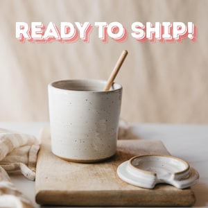 May include: A speckled white ceramic mug with a wooden spoon inside, resting on a wooden cutting board. The mug has a matching lid with a spoon rest. The text "READY TO SHIP!" is displayed above the mug.