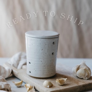 May include: A speckled, off-white ceramic garlic keeper with a lid, featuring several small holes for ventilation. The keeper sits on a wooden cutting board with garlic cloves and a whole bulb. The text "READY TO SHIP" is above the keeper.