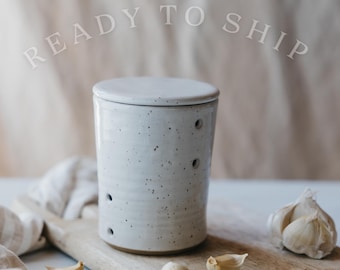 May include: A speckled, off-white ceramic garlic keeper with a lid, featuring several small holes for ventilation. The keeper sits on a wooden cutting board with garlic cloves and a whole bulb. The text "READY TO SHIP" is above the keeper.